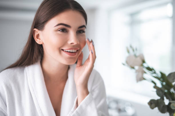 Young woman with smooth, glowing skin looking in the mirror after Hyperdilute Radiesse collagen treatment at a medspa